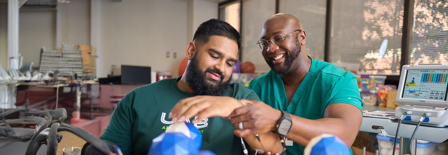 Two men, one in green scrubs and the other in a UAB T-shirt, smile as they work together with medical equipment in a brightly lit room, possibly engaging in a healthcare or educational activity.