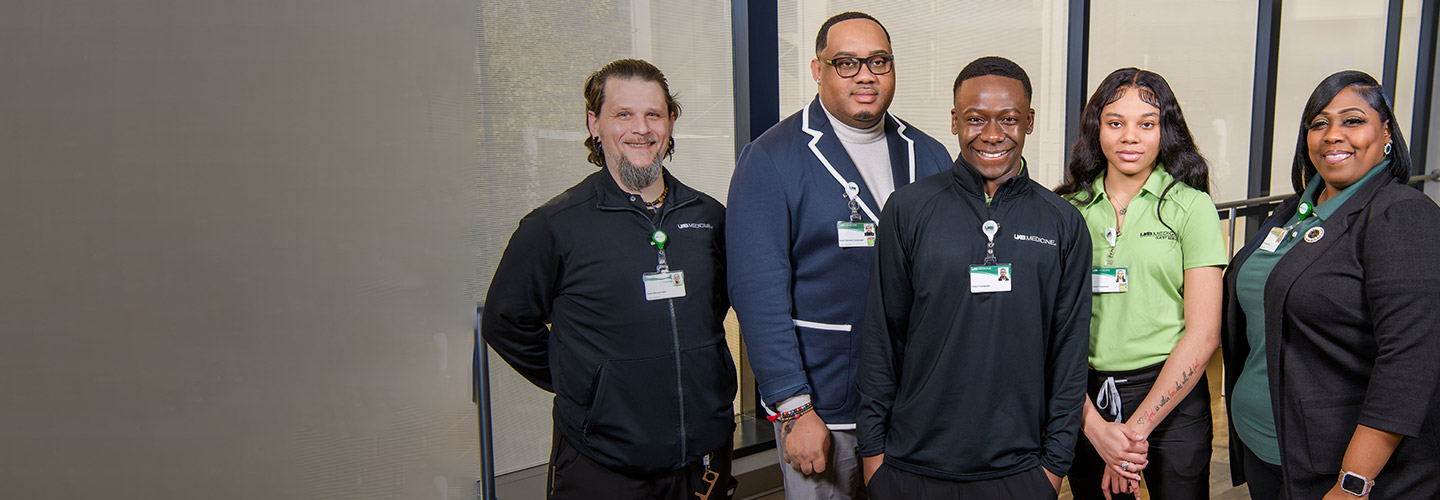 Five professionally dressed people, three men and two women, stand indoors smiling at the camera. Four wear lanyards and name badges, and one woman wears a green shirt while the others wear black or dark clothing.