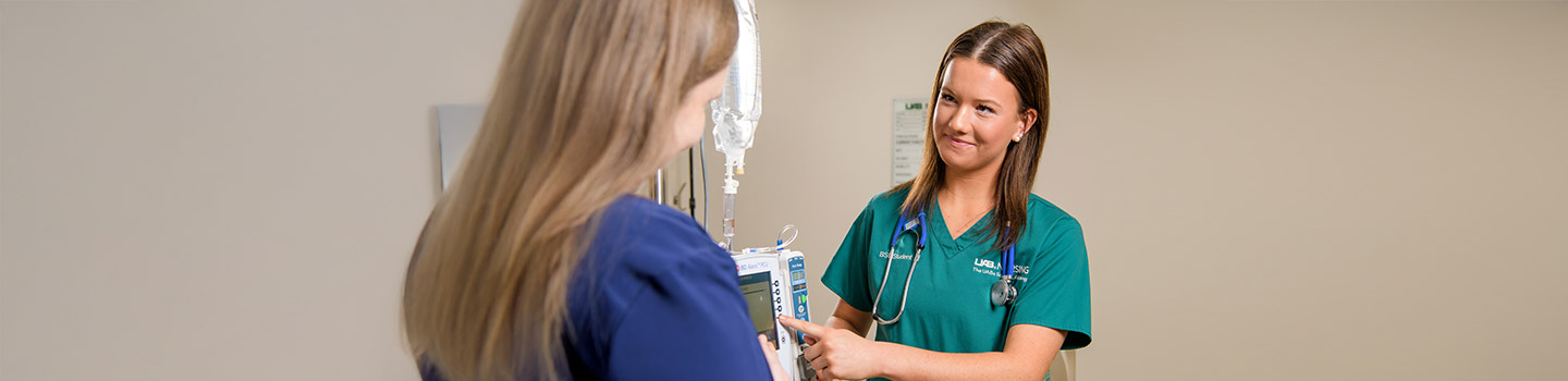 A nurse in green scrubs stands beside a patient, adjusting an IV drip and infusion pump, while smiling and making eye contact in a medical setting.