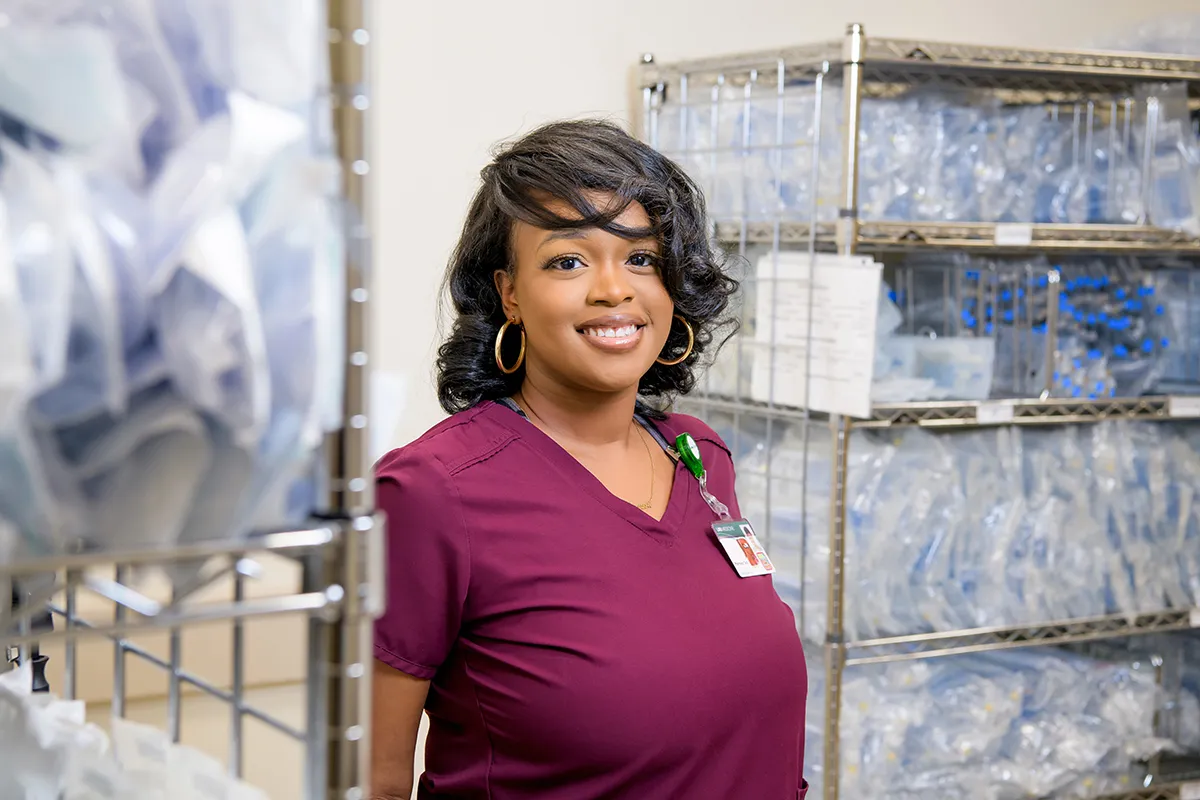 A woman in burgundy scrubs smiles while standing in a medical supply room filled with organized shelves of packaged medical equipment. She wears a badge and hoop earrings.
