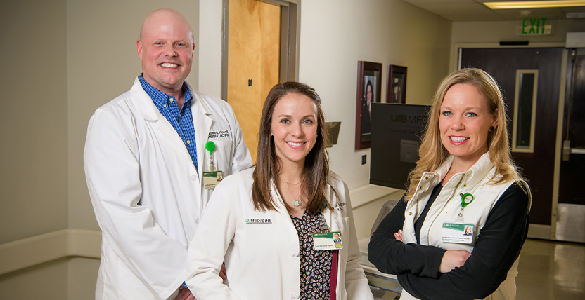 Three smiling medical professionals, two women and one man, stand in a hallway wearing white coats and name badges. They appear confident and approachable, with medical equipment visible in the background.