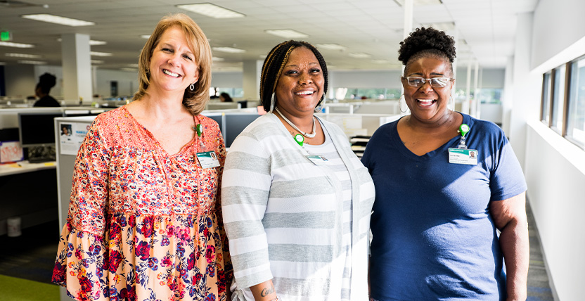Three women stand together in a brightly lit office, smiling at the camera. They all wear employee badges and are dressed in casual, professional attire. Cubicles and large windows are visible in the background.
