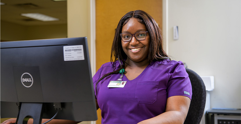 A woman in purple scrubs sits at a desk, smiling at the camera. She wears glasses and a name badge, and is positioned in front of a computer monitor in an office setting.