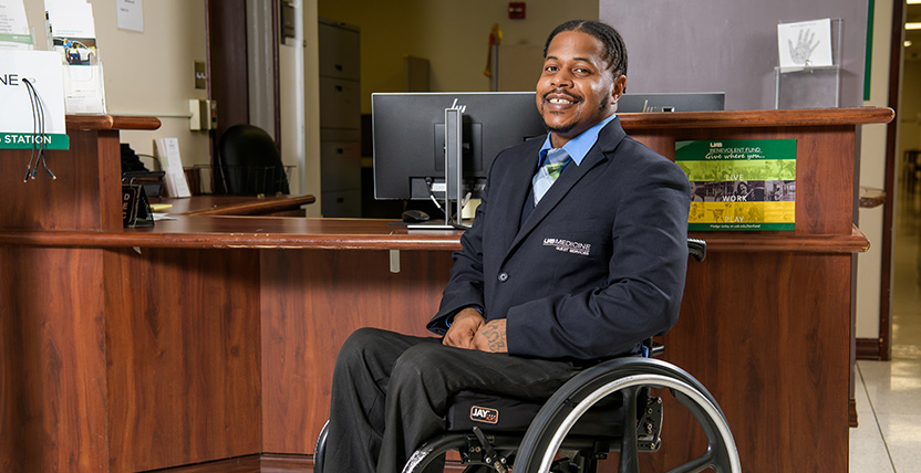A smiling man in a suit sits in a wheelchair in front of a reception desk, with office equipment and signage in the background.