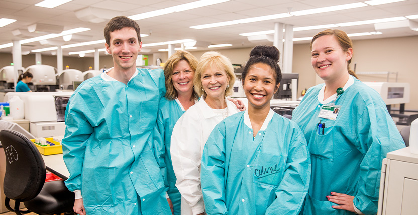 Five people, including three in teal lab coats and two in white coats, stand together smiling in a brightly lit laboratory with medical equipment in the background.