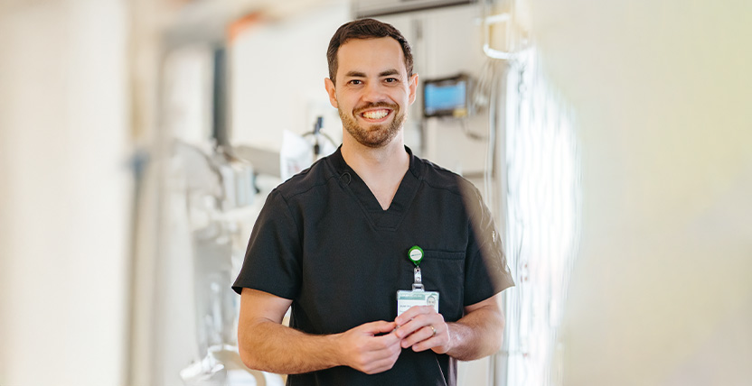 A smiling healthcare professional wearing black scrubs and a name badge stands in a brightly lit medical facility hallway.