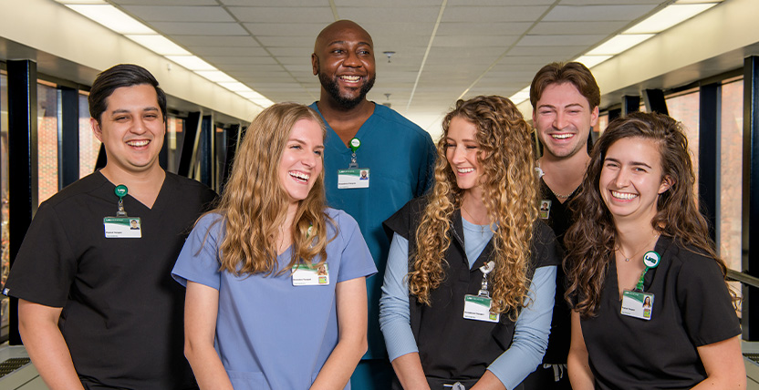 Six healthcare professionals wearing scrubs and name badges stand together in a hallway, smiling and laughing. The background shows a bright corridor with large windows.