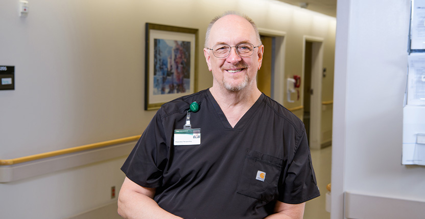 A smiling male healthcare professional wearing black scrubs and an ID badge stands in a well-lit hallway of a medical facility. A framed picture and medical equipment are visible in the background.