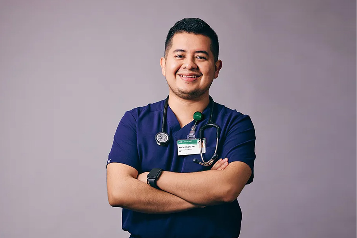A smiling healthcare professional wearing navy blue scrubs, a stethoscope, and a name badge stands with arms crossed against a plain background.