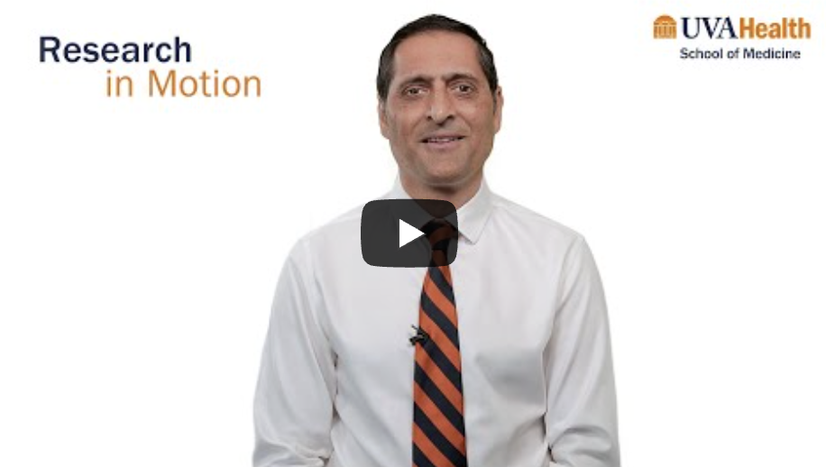 A person wearing a white dress shirt and an orange‑and‑navy striped tie sits centered against a white background. A play button icon appears over the chest area, indicating a video. The text ‘Research in Motion’ is on the left, and the UVA Health School of Medicine logo is on the upper right..